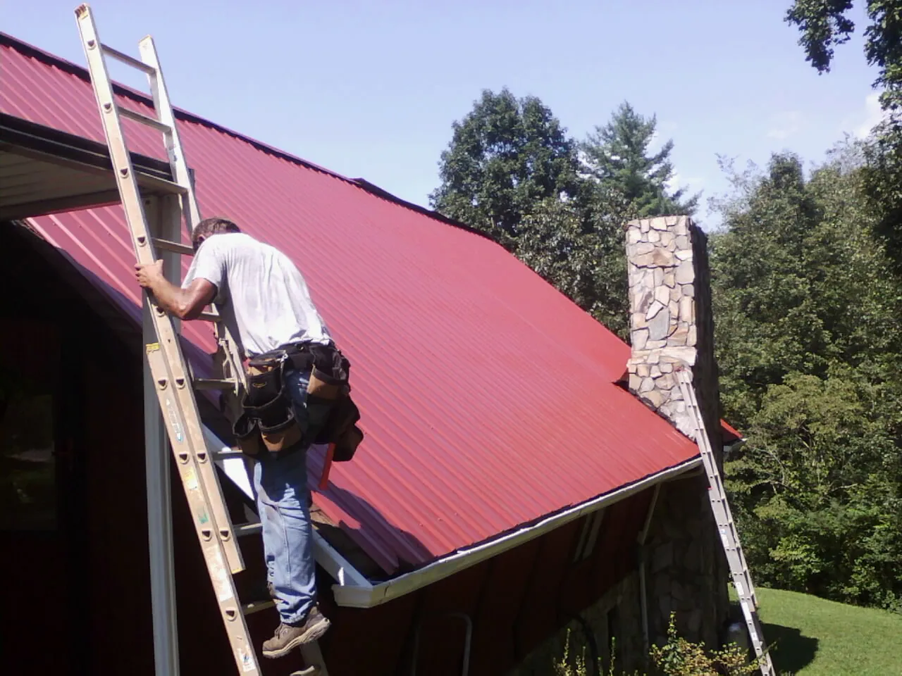 Roofer on a ladder beside a red metal roof and stone chimney — Soffit & Fascia Repair by Roofing Brothers in Riner